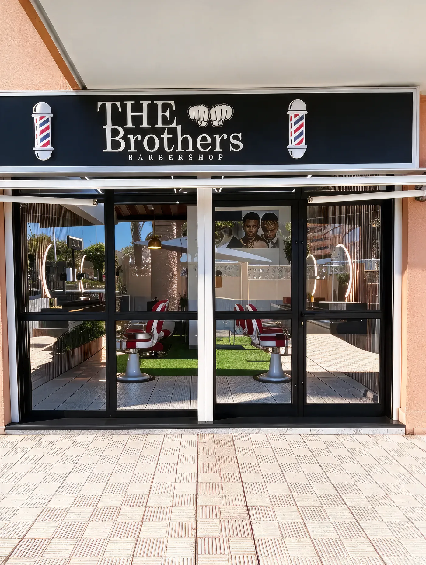 Fachada de The Brothers Barbershop: letrero negro con los puños y dos postes de barbero, escaparate de cristal con las sillas rojas dentro